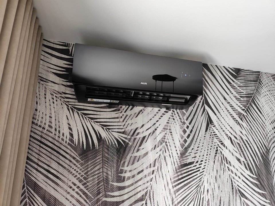 Czarny klimatyzator marki AUX zamontowany na ścianie z tapetą w motywy liści palmowych, widoczny fragment zasłony po lewej stronie.