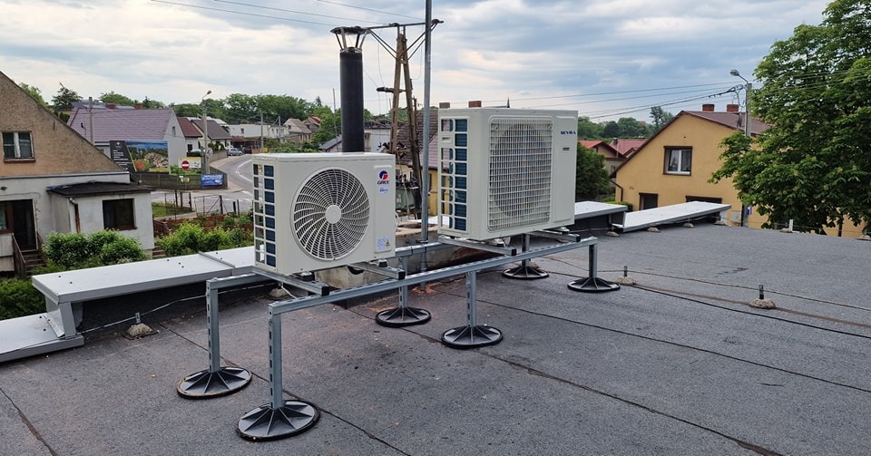Dwa białe klimatyzatory marki Gree i Seyra zamontowane na metalowej konstrukcji na płaskim, bitumicznym dachu budynku mieszkalnego, widok z góry na tle zabudowy jednorodzinnej i pochmurnego nieba.
