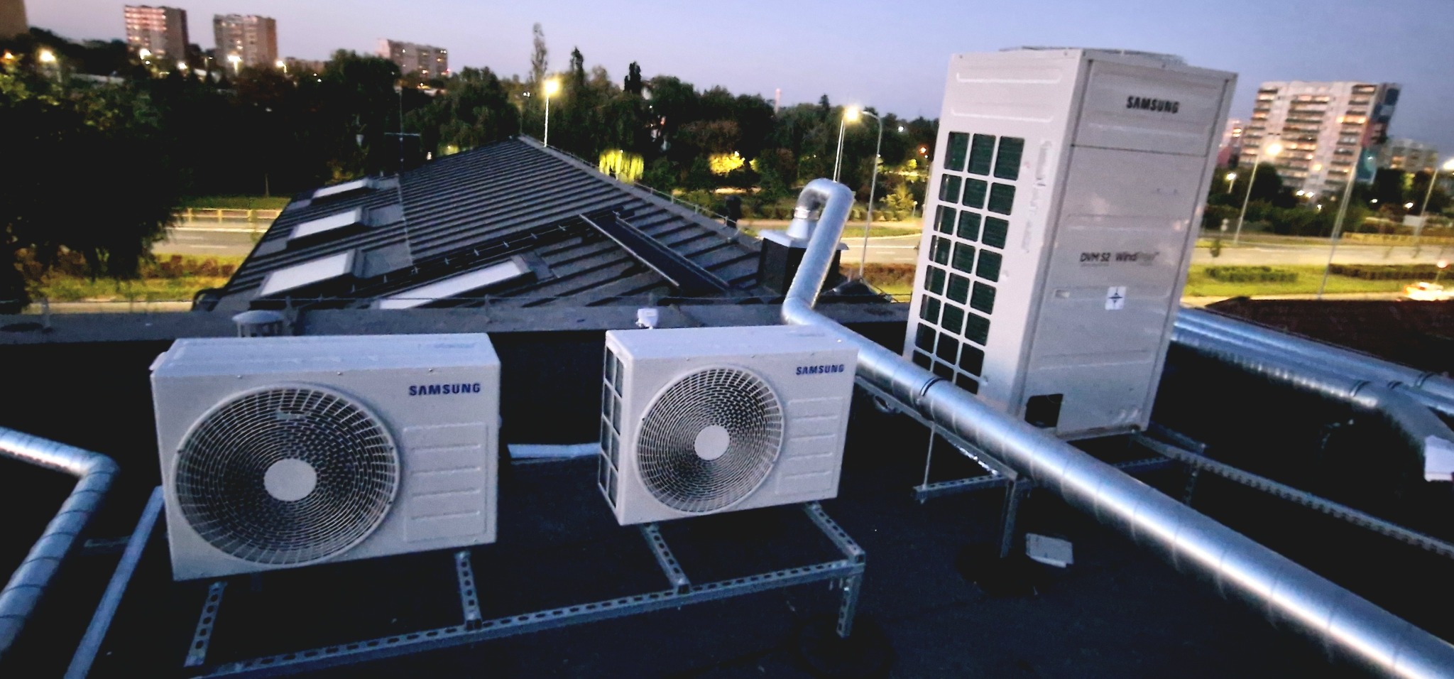Instalacja zewnętrznych jednostek klimatyzacyjnych Samsung na dachu budynku z widokiem na miasto o zmierzchu, rury instalacyjne w srebrnym kolorze.