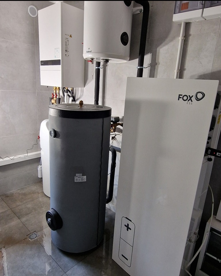 Szary akumulator ciepła z podłączonymi rurami, biały zasobnik ciepłej wody i biała jednostka magazynowania energii FOX ESS w kotłowni w Koninie, widoczne szare płytki na ścianach i podłodze.
