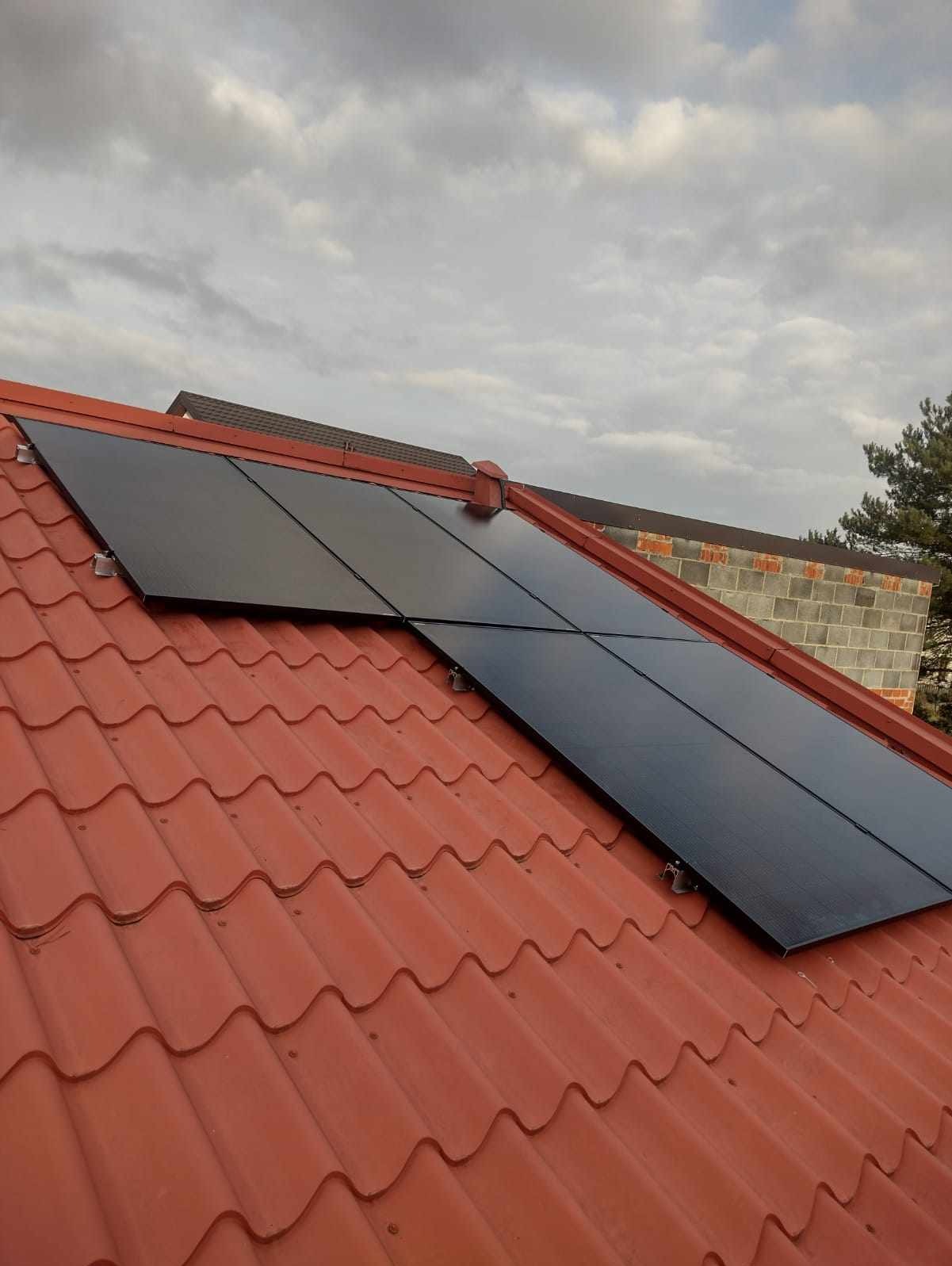 Panele słoneczne zamontowane na dachu z czerwonej dachówki, widoczny komin z cegły i pochmurne niebo w tle.