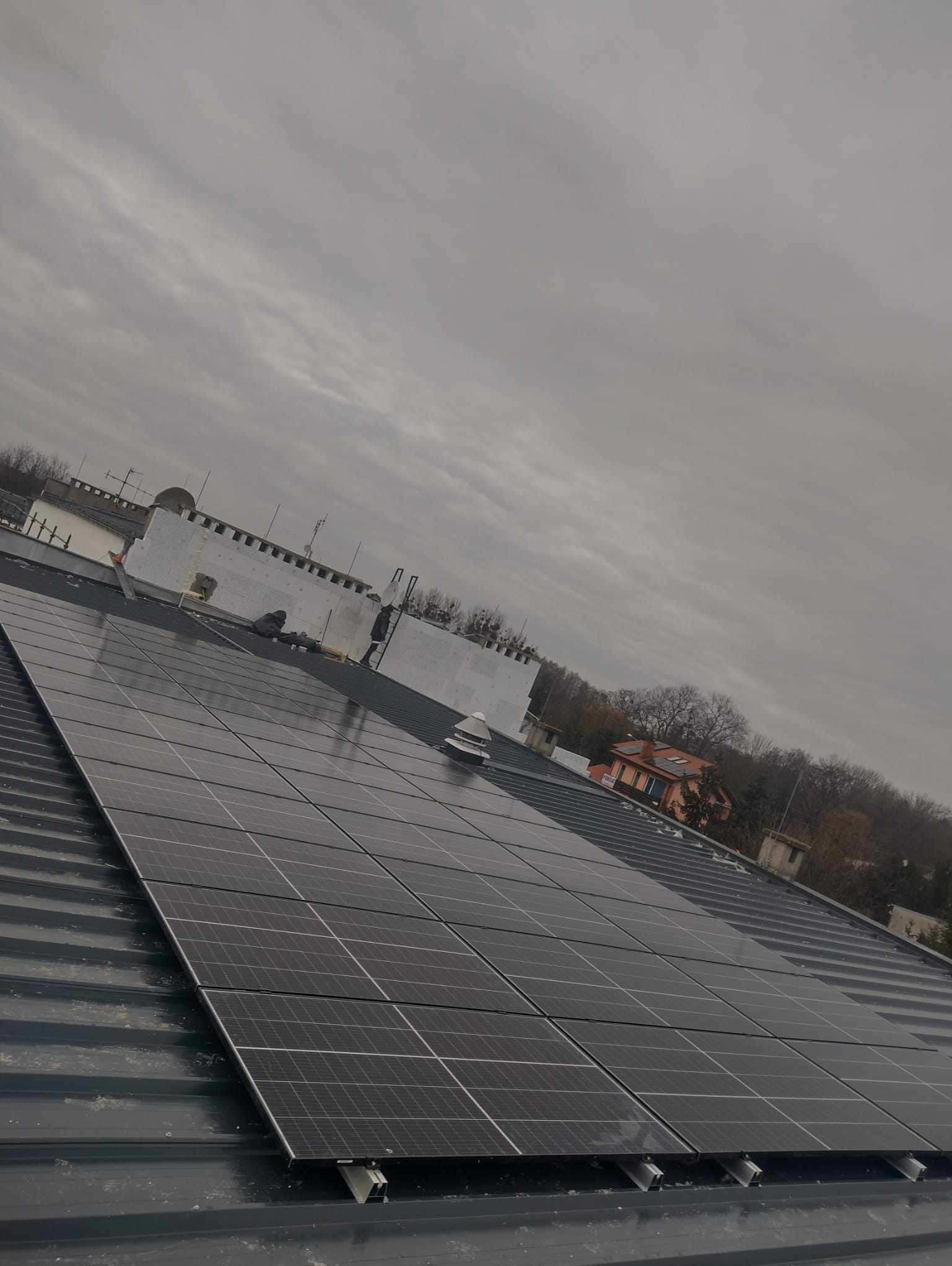 Instalacja paneli słonecznych na metalowym dachu w pochmurny dzień, widoczne elementy montażowe i budynek w tle.