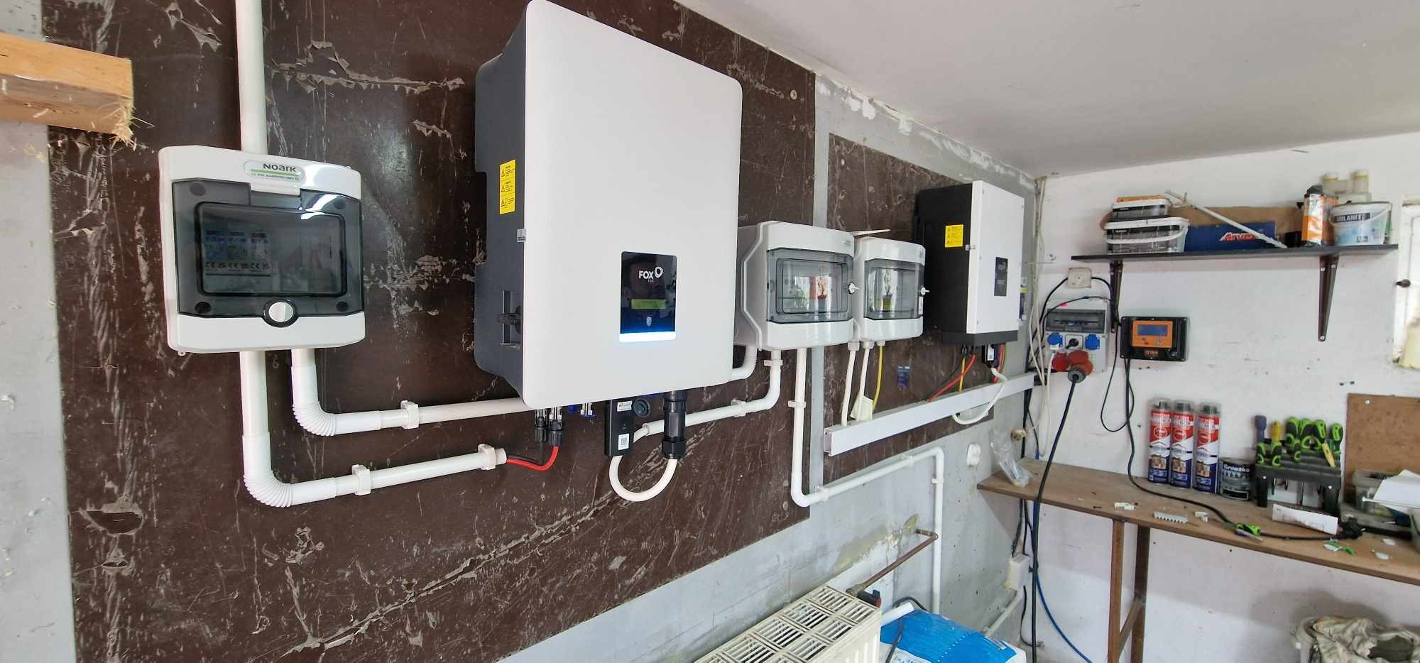 Instalacja fotowoltaiczna w pomieszczeniu: biały inwerter FOXO, rozdzielnica NOARK i zabezpieczenia na ścianie z brązową okładziną, widoczne przewody i elementy montażowe.