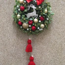 Dekoracyjny wieniec świąteczny z zielonych liści, czerwonych jabłek, złotych i białych bombek, białego renifera w środku, zwieńczony czerwoną kokardą i ozdobiony czerwonymi frędzlami.