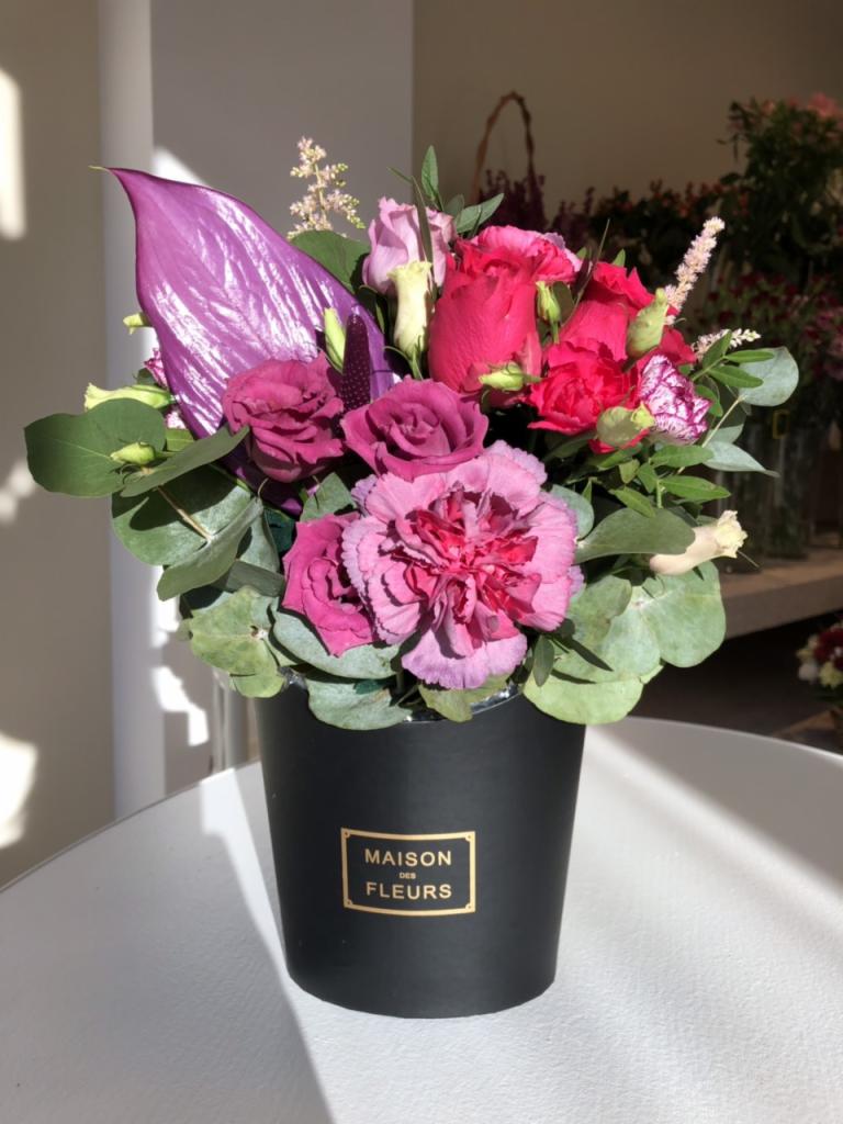 Kompozycja kwiatowa w czarnym pudełku z napisem 'Maison des Fleurs', zawierająca różowe róże, goździki i anturium w odcieniach fioletu, na jasnym blacie.