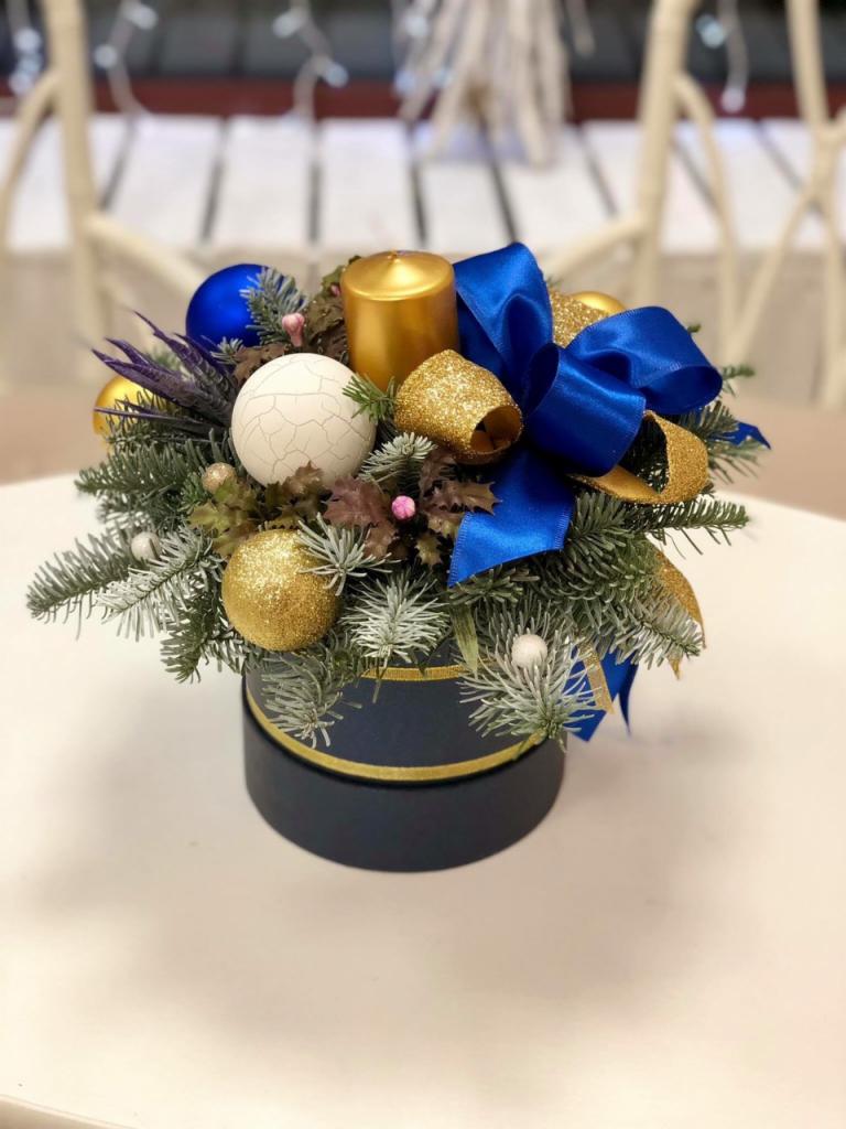 Kompozycja świąteczna w okrągłym, granatowym pudełku, z zielonymi gałązkami, złotą świecą, niebieską kokardą i ozdobami w kolorach złota, bieli i niebieskiego, umieszczona na białym stole.