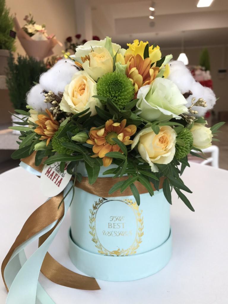 Kompozycja kwiatowa w pastelowo-błękitnym pudełku z napisem 'The Best Wishes', zawiera róże w odcieniach żółci i bieli, pomarańczowe chryzantemy, zielone kule, gałązki bawełny oraz wstążki...