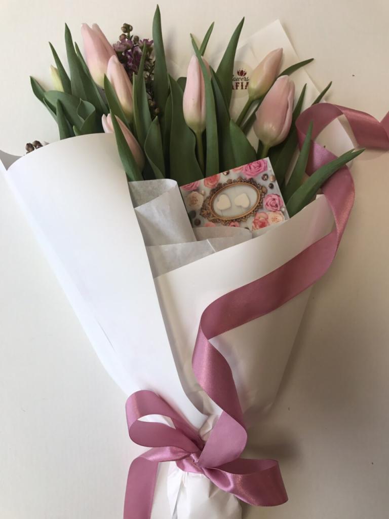 Bukiet jasnoróżowych tulipanów zawinięty w biały papier, przewiązany satynową wstążką w odcieniu pudrowego różu, z dołączonym bilecikiem z motywem serc i róż