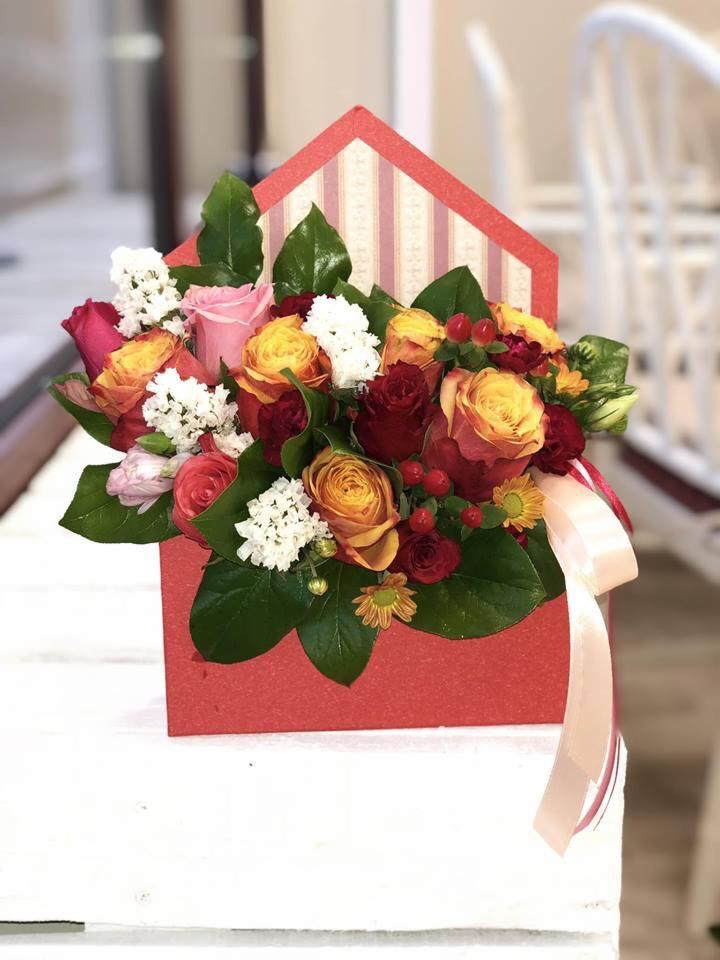 Kompozycja kwiatowa z róż, goździków i drobnych białych kwiatów w czerwonym pudełku w kształcie domu z beżowo-białymi paskami, przewiązana beżową wstążką.