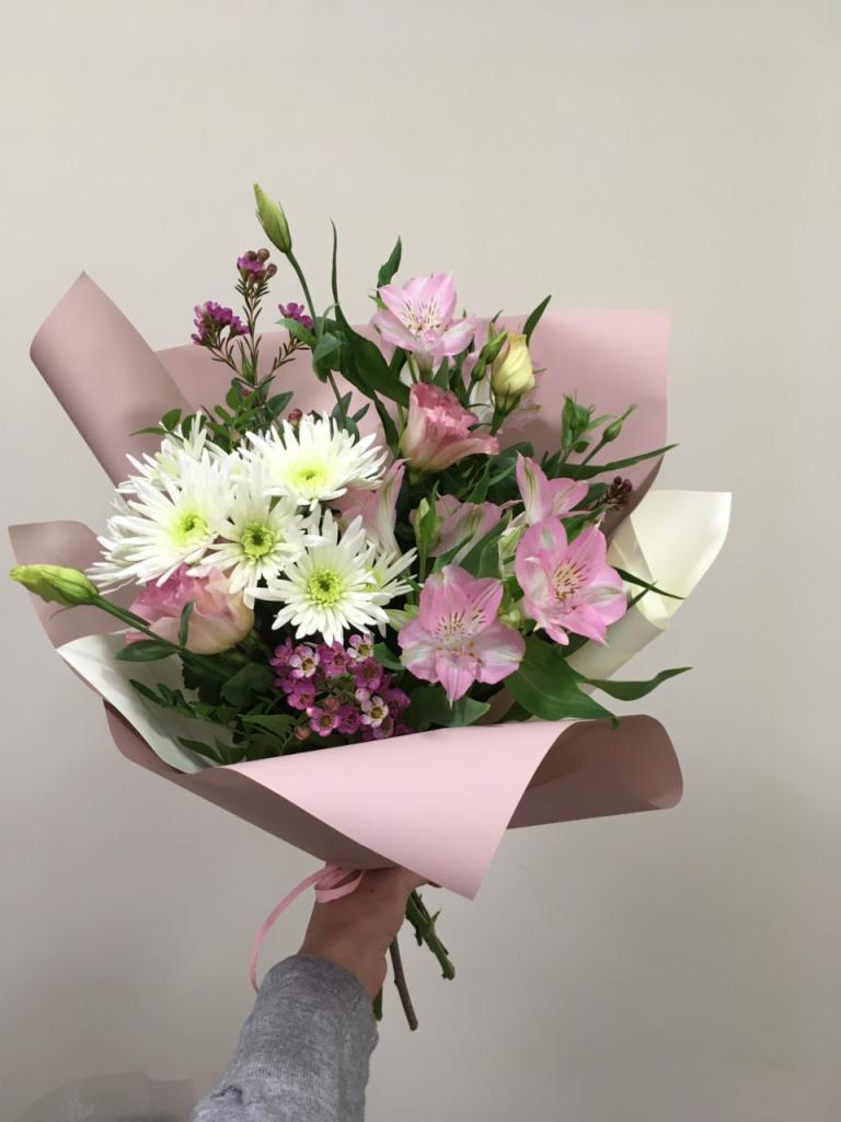 Bukiet mieszanych kwiatów w pastelowym różowym papierze, trzymany w dłoni, z widocznymi białymi chryzantemami, różowymi alstromeriami i drobnymi fioletowymi kwiatami.