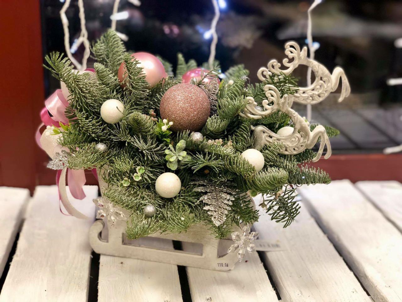 Świąteczna kompozycja kwiatowa w drewnianych sankach, z gałązkami świerku, różowymi i białymi bombkami, dekoracyjnymi reniferami i kokardą, na białym drewnianym stole.
