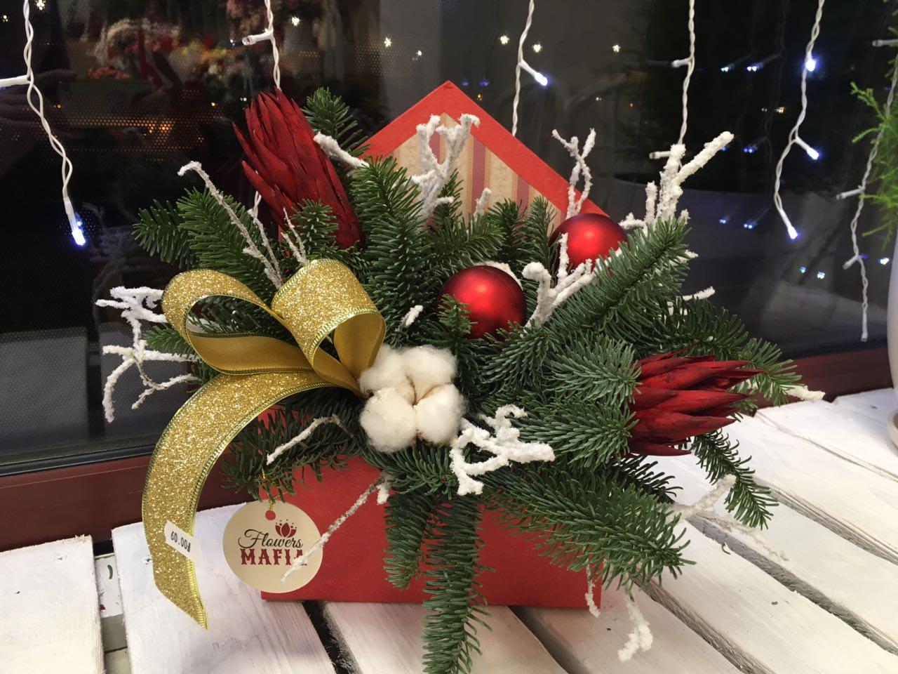 Kompozycja świąteczna w czerwonym pudełku z gałązkami świerku, czerwonymi bombkami, suszem, bawełną i złotą, brokatową wstążką, z zawieszką z logo 'Flowers Mafia'.