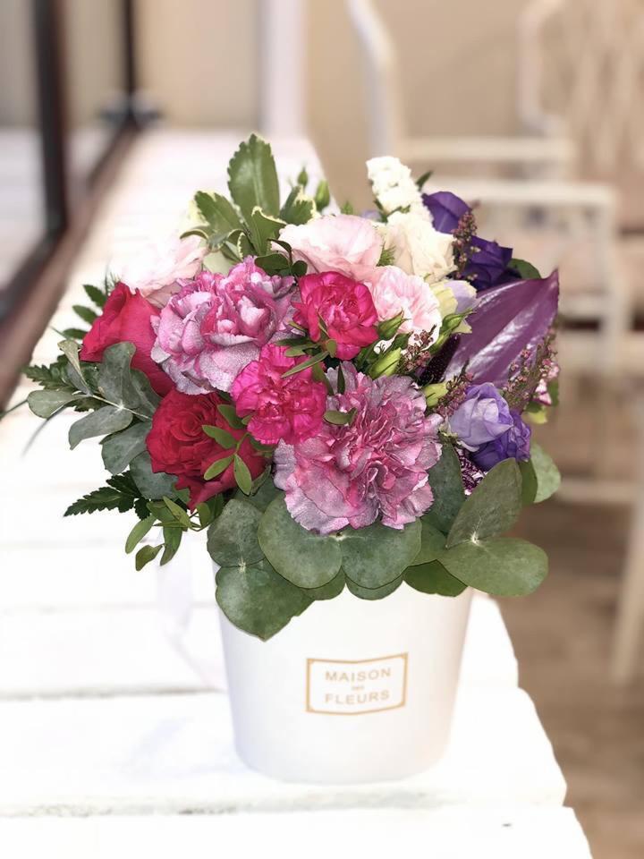 Kompozycja kwiatowa w białym pudełku z napisem 'Maison des Fleurs', zawierająca różowe goździki, czerwone róże, białe eustomy, fioletowe kwiaty i zielone liście eukaliptusa.