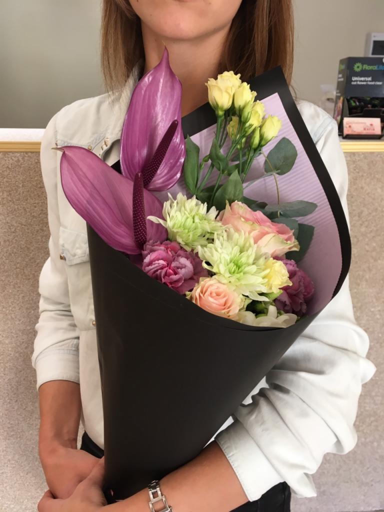 Kobieta trzyma bukiet mieszanych kwiatów w czarnym papierze, z widocznym anturium, różami, goździkami i eustomą, delikatne pastelowe kolory.