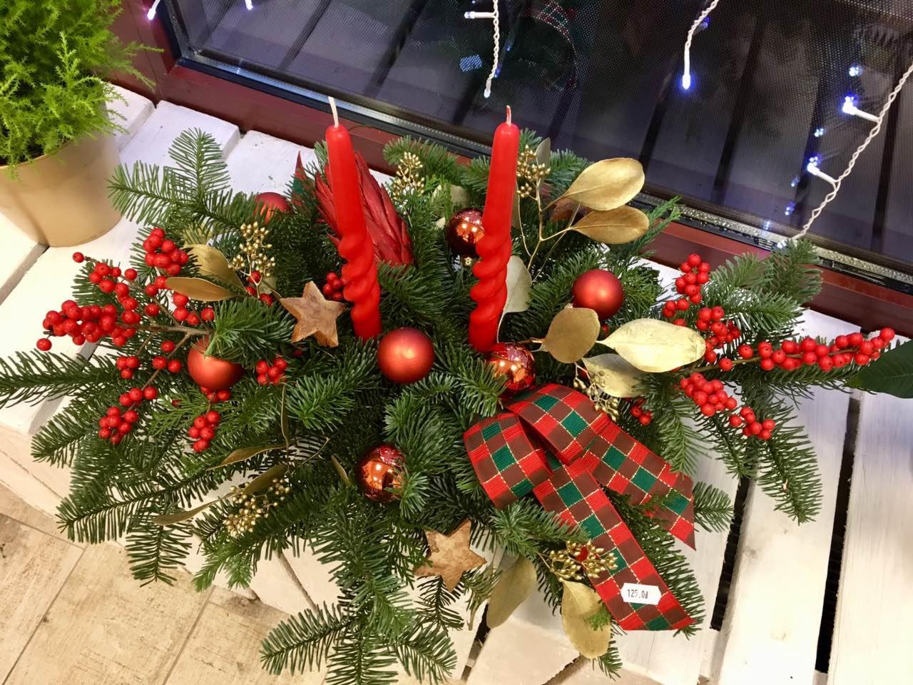 Dekoracja świąteczna z gałązek choinki, czerwonych świec, bombek, złotych liści i czerwono-zielonej wstążki w kratę, umieszczona na białym parapecie okna z widocznymi lampkami.