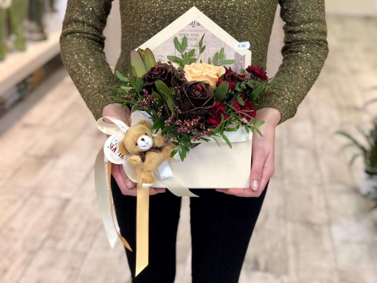 Kobieta trzyma kompozycję kwiatową w pudełku w kształcie koperty z pluszowym misiem, zawierającą ciemnobrązowe, czerwone i kremowe róże oraz zielone dodatki.