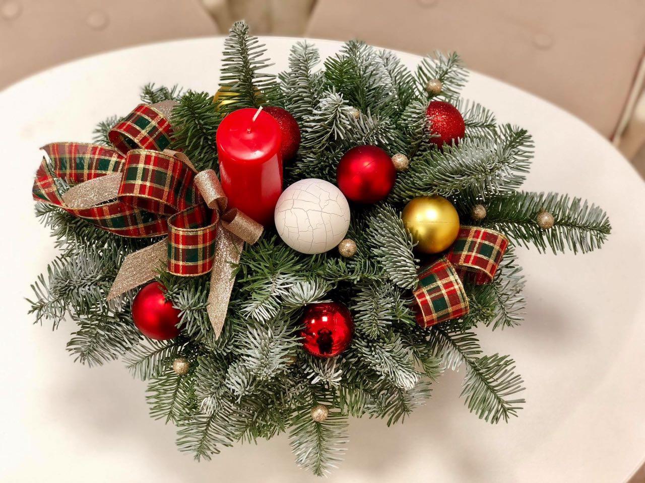 Świąteczna kompozycja stołowa z gałązek świerkowych oprószonych sztucznym śniegiem, czerwonej świecy, czerwonych i złotych bombek oraz wstążek w kratę i złotych kokardek na białym stole.