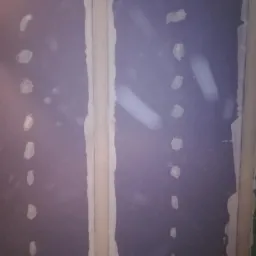 Fragment ściany z płyt kartonowo-gipsowych, na której widoczne są miejsca po wkrętach pokryte szpachlą, gotowe do dalszej obróbki.