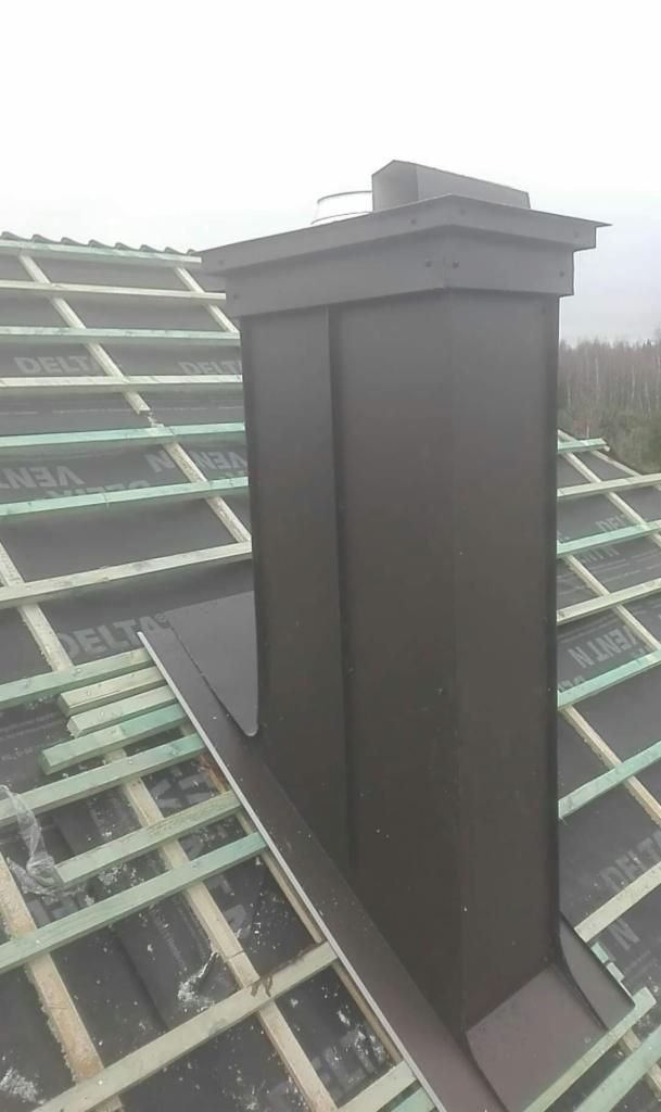 Czarny, metalowy komin na dachu w trakcie budowy, widoczne drewniane elementy konstrukcji dachu i membrana dachowa.