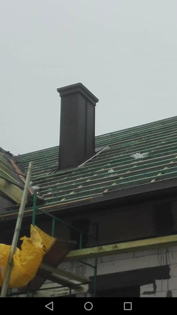 Widok dachu w trakcie budowy z widocznym kominem, łatami pod pokrycie i częściowo zadaszoną konstrukcją, z rusztowaniem i żółtą folią ochronną w tle.