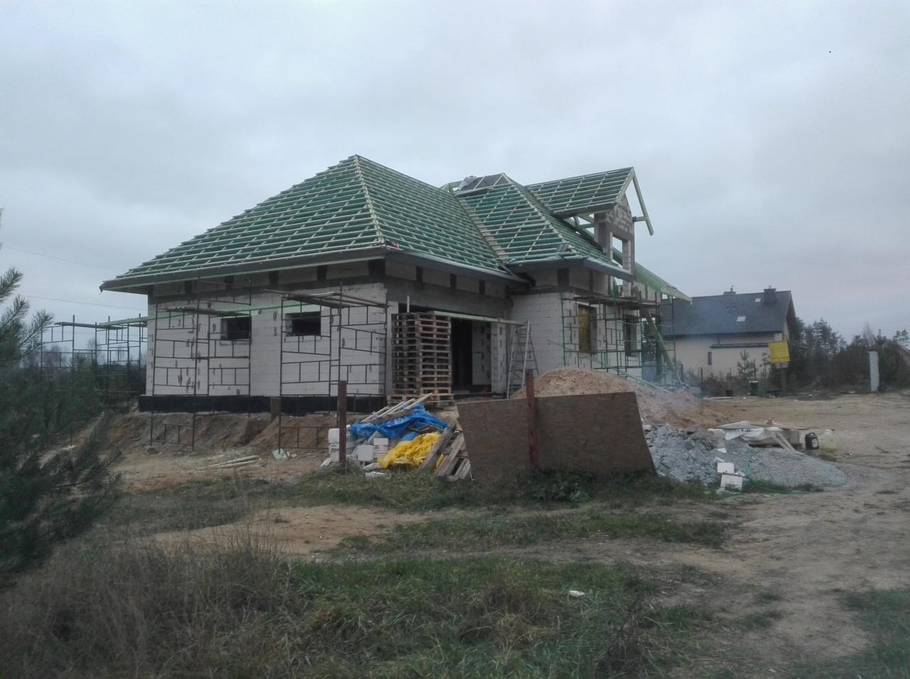 Dom w trakcie budowy z widoczną więźbą dachową i zielonym deskowaniem pod pokrycie, palety i materiały budowlane na działce.