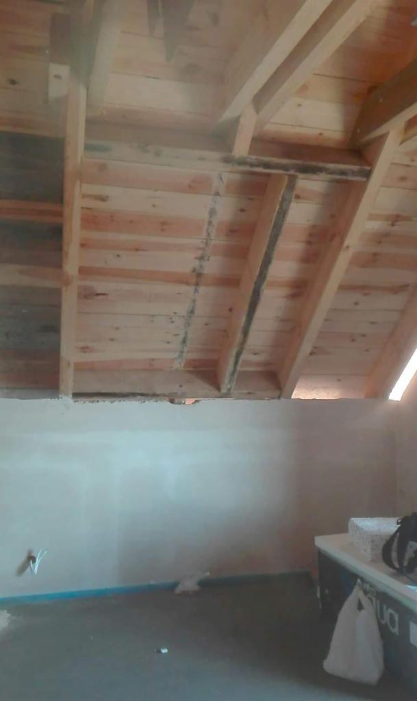 Surowe drewno więźby dachowej w trakcie montażu, widoczne elementy konstrukcyjne i odeskowanie. Ściana w tle częściowo otynkowana, widoczne ślady prac budowlanych.