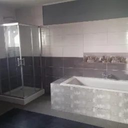 Wykończona łazienka w Elblągu z kabiną prysznicową, wanną obudowaną płytkami z motywem kwiatowym oraz dwukolorowymi płytkami na ścianach z motywem miasta.