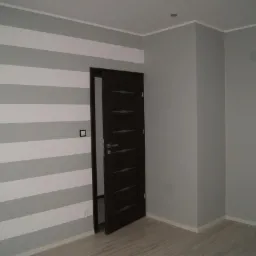 Pokój po świeżym malowaniu z szarymi i białymi pasami na ścianie, widoczne gniazdka elektryczne i włącznik światła, otwarte ciemne drzwi z poziomymi przeszkleniami.