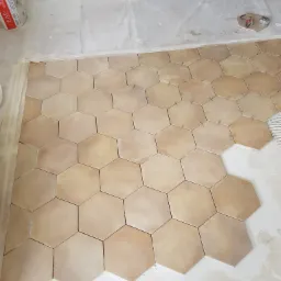 Układanie beżowych, heksagonalnych płytek podłogowych z użyciem krzyżyków dystansowych, widoczny fragment podłogi z częściowo ułożonymi płytkami i paczka płytek w tle.