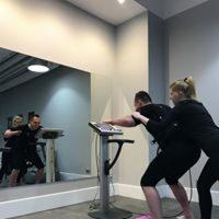 Trening EMS w Warszawie: kobieta w czarnym stroju pomaga mężczyźnie w czarnym stroju ćwiczyć z wykorzystaniem specjalistycznego urządzenia do elektrostymulacji mięśni, widoczne odbicie w lustrze.