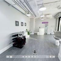 Jasne, przestronne wnętrze siłowni lub studia fitness z szarymi podłogami, białymi ścianami, widocznymi instalacjami na suficie, rośliną doniczkową i sprzętem do ćwiczeń na stojaku.