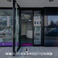 Wejście do jasnego, minimalistycznego studia z recepcją widoczną w tle. Otwarta szklana witryna z czarnymi ramami, widoczny fragment chodnika i odbicie ulicy w szybach.