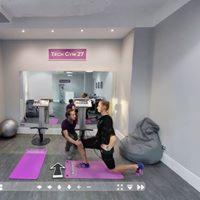 Osoba w czarnym stroju sportowym wykonuje wykrok na fioletowej macie w jasnym pomieszczeniu z lustrem i napisem 'Trio Gym 27'. Obok stoi dziecko, obserwując ćwiczenie.