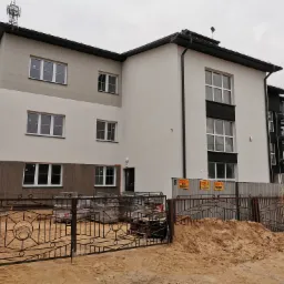 Nowo wybudowany, trzypiętrowy budynek z anteną na dachu, ogrodzony metalowym płotem, z widocznymi materiałami budowlanymi i rusztowaniami w tle.