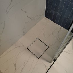 LUXBUD - Nowoczesna kabina prysznicowa z odpływem liniowym, wyłożona białymi płytkami imitującymi marmur na podłodze i ścianie, oraz granatowymi, pionowo ułożonymi płytkami na bocznej ścianie.