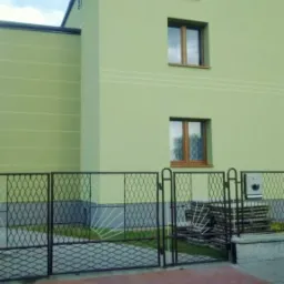 Jasnozielony dom z nową elewacją, widoczna metalowa brama z dekoracyjnym wzorem, stos płytek za bramą, trzy okna z brązowymi ramami.
