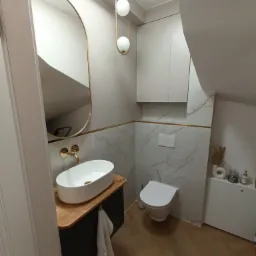 Nowoczesna łazienka na poddaszu z owalną umywalką nablatową, złotą armaturą i lustrem w złotej ramie, marmurowe płytki ścienne z dekoracyjną złotą listwą, wisząca toaleta i drewnopodobna podłoga...