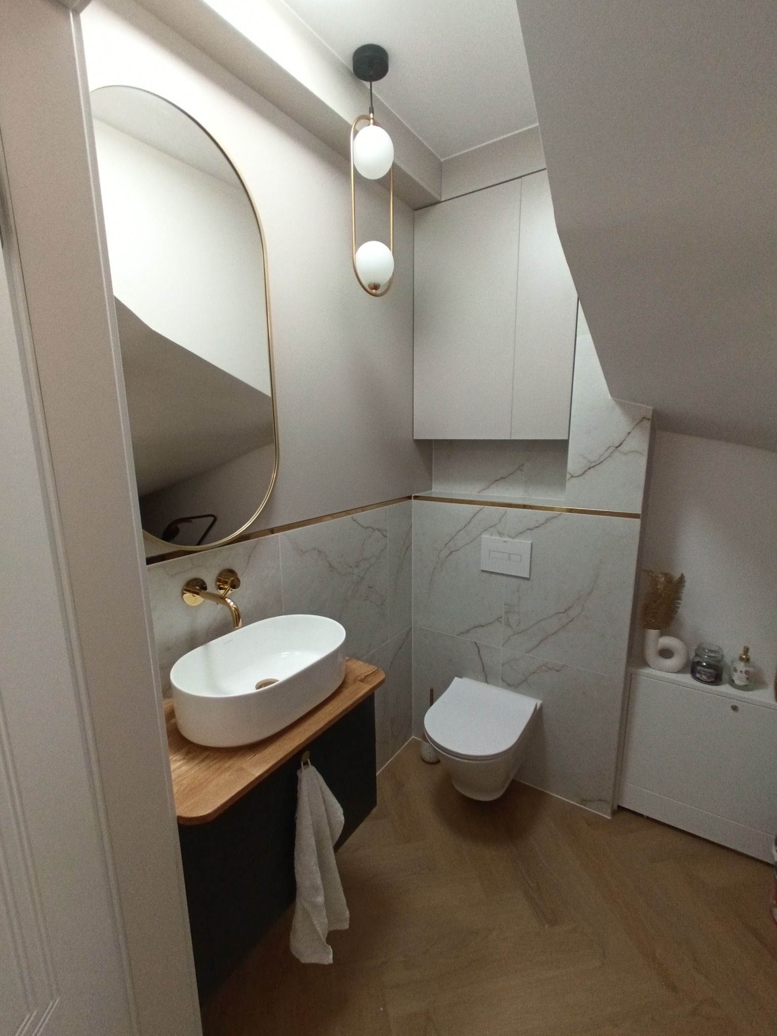 Nowoczesna łazienka na poddaszu z owalną umywalką nablatową, złotą armaturą i lustrem w złotej ramie, marmurowe płytki ścienne z dekoracyjną złotą listwą, wisząca toaleta i drewnopodobna podłoga...