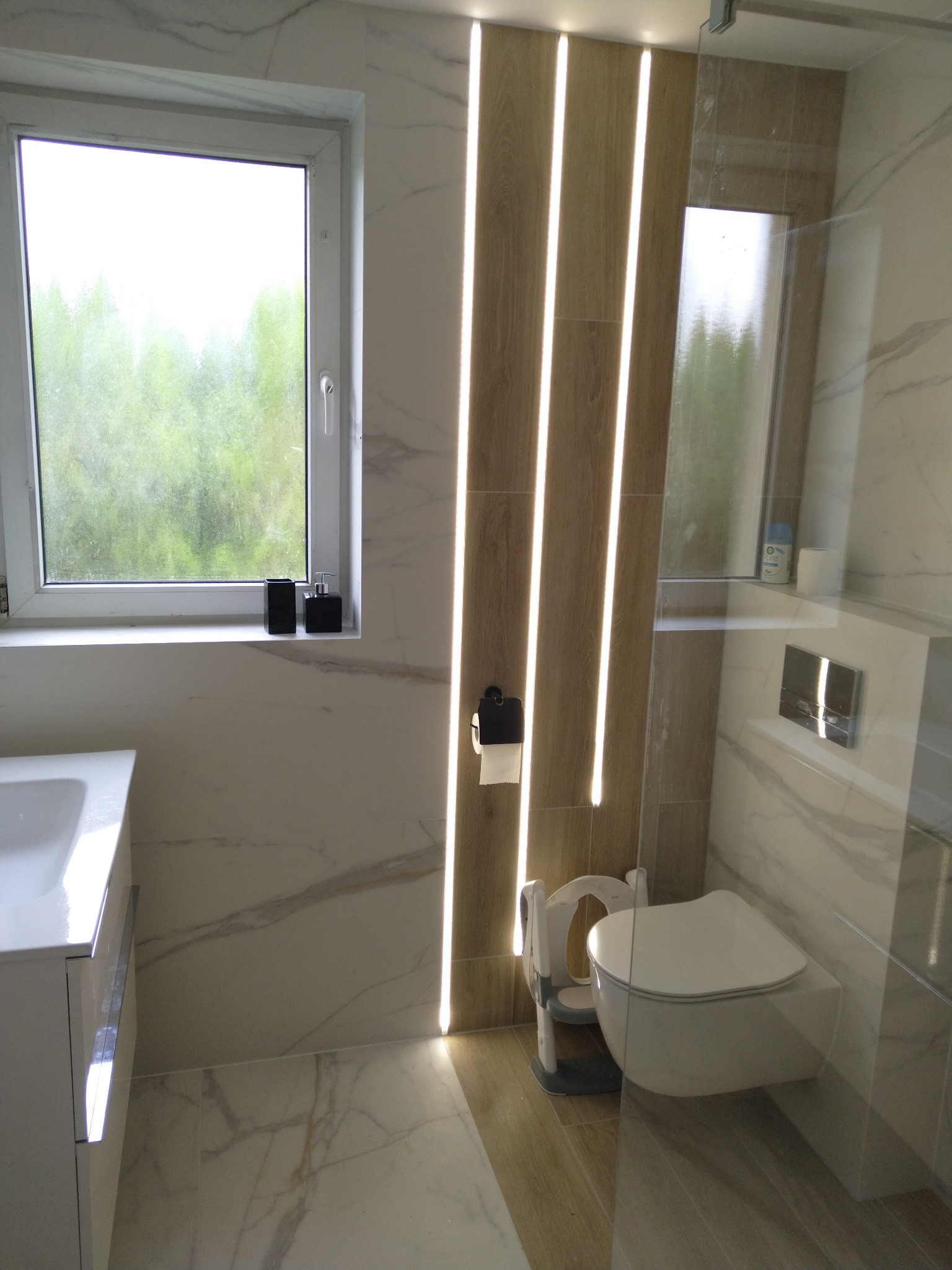 Nowoczesna łazienka z marmurowymi płytkami, podwieszaną toaletą z nakładką dla dziecka, oświetleniem LED w pionowych panelach i oknem z widokiem na zieleń.
