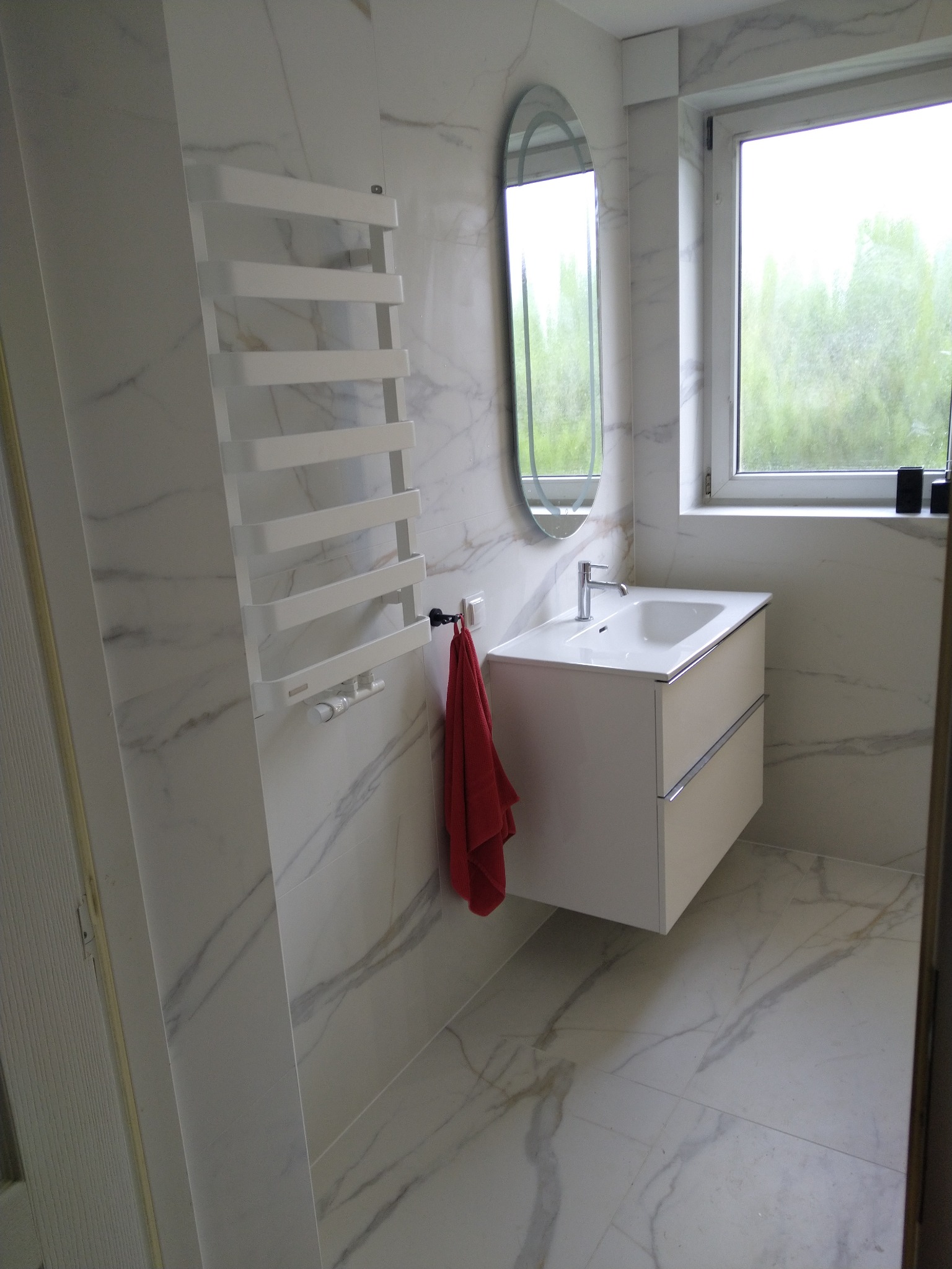 Nowoczesna łazienka z białymi płytkami imitującymi marmur, owalne lustro, biała szafka pod umywalką z dwiema szufladami, grzejnik drabinkowy i czerwony ręcznik