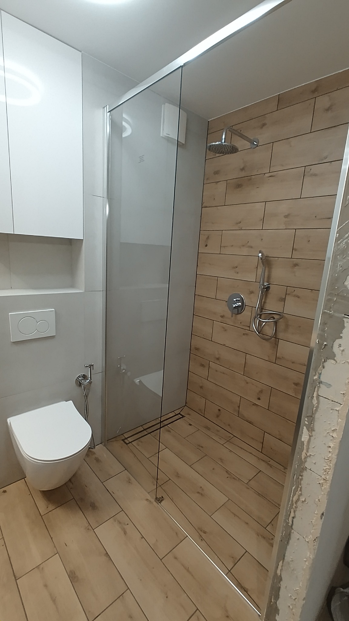 Nowoczesna łazienka z kabiną prysznicową typu walk-in, wykończona płytkami imitującymi drewno, z podtynkową armaturą i wiszącą miską WC.