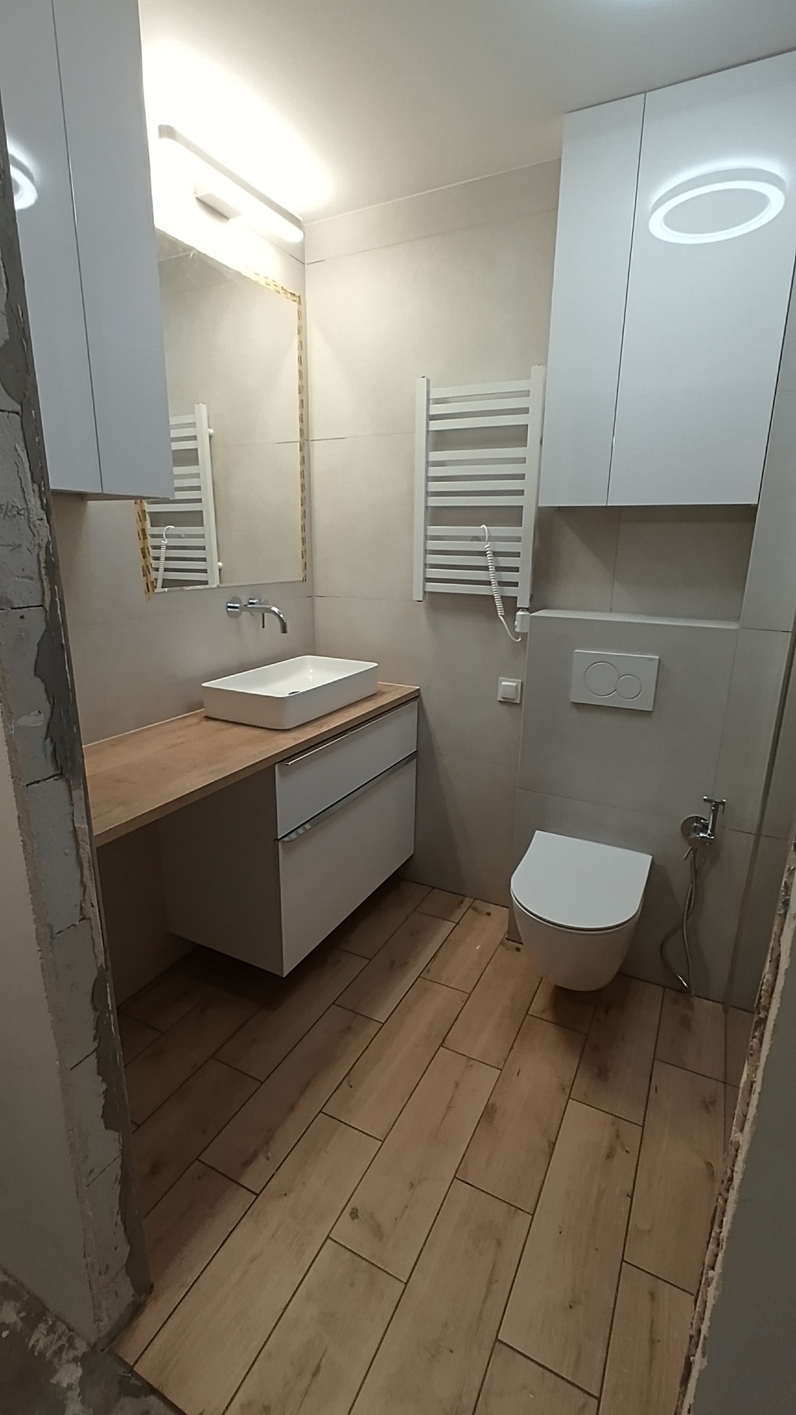 Wykończona łazienka z drewnopodobną podłogą, białą umywalką nablatową na drewnianym blacie, wiszącą toaletą i grzejnikiem drabinkowym, oświetlona lampą LED na szafce.