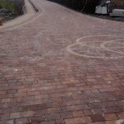 Długa, kręta droga z czerwonej kostki brukowej z dekoracyjnym wzorem w kształcie koła, prowadząca do bramy wjazdowej z widocznym pomarańczowym pojazdem i żywopłotem w tle, pochmurny dzień.