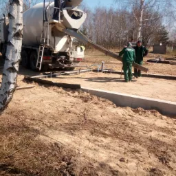 Betonomieszarka wylewa beton do przygotowanego szalunku fundamentowego; pracownicy w zielonych kombinezonach nadzorują proces; w tle drzewa bez liści i błękitne niebo.