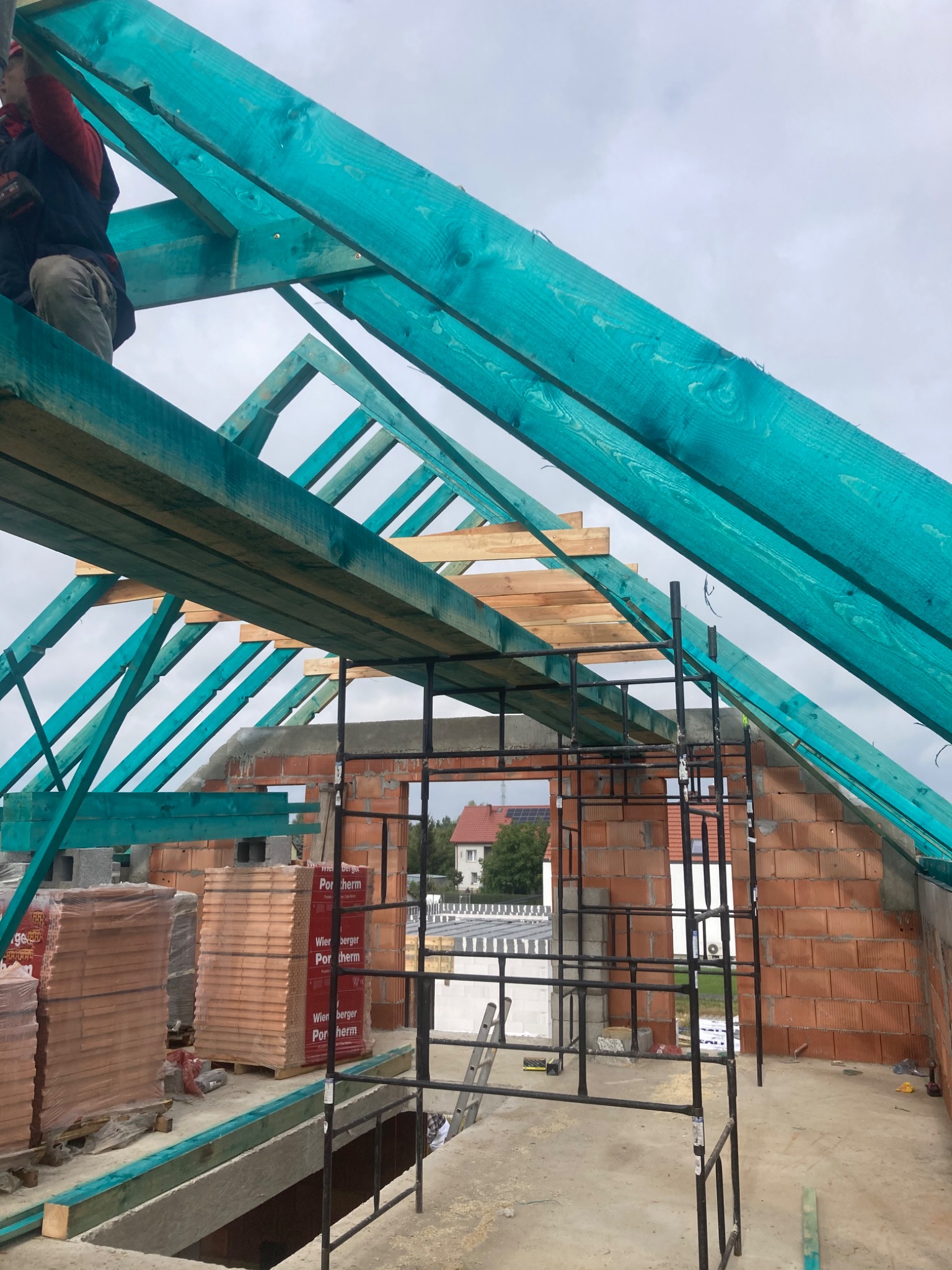 Wnętrze surowego domu w trakcie budowy: widoczne czerwone cegły ścian, rusztowanie, drewniana konstrukcja dachu pomalowana na turkusowo, palety z cegłami, oraz pracownik na dachu.