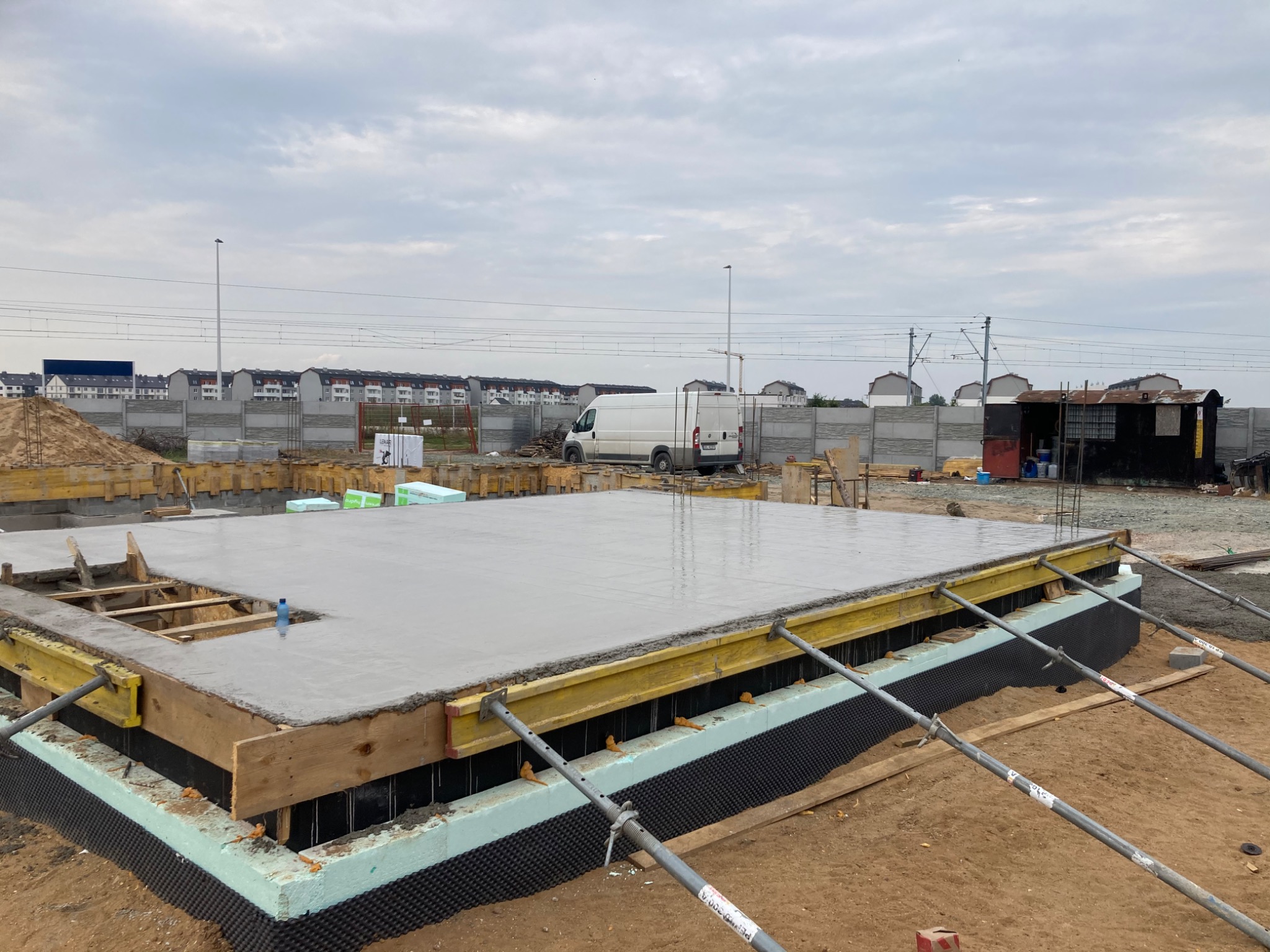 Wylana płyta fundamentowa z deskowaniem i izolacją termiczną, widoczne stalowe podpory i butelka wody, w tle zabudowa wielorodzinna i linia kolejowa.