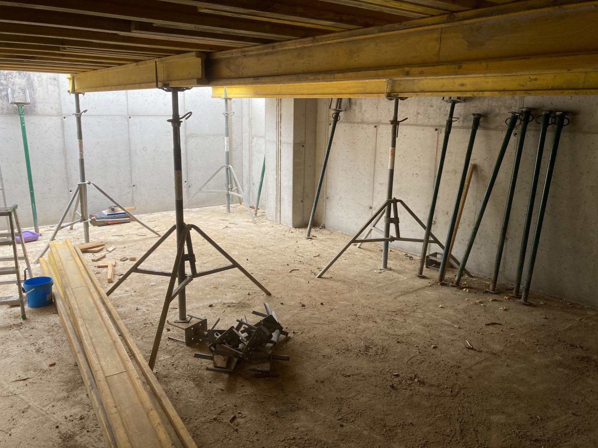 Wnętrze w trakcie budowy z szalunkami stropowymi podpartymi stalowymi stemplami, widoczne betonowe ściany i podłoże, elementy szalunków leżące na ziemi.