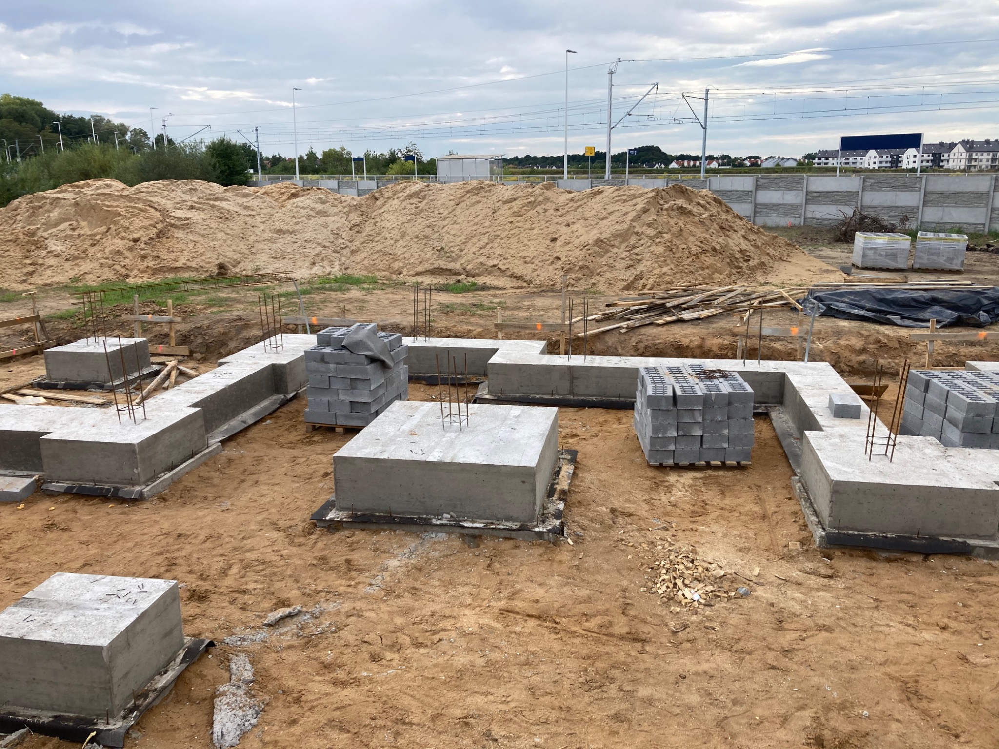 Fundamenty pod budynek w trakcie wylewania, widoczne zbrojenia i bloczki betonowe, w tle hałda piasku i linia kolejowa.