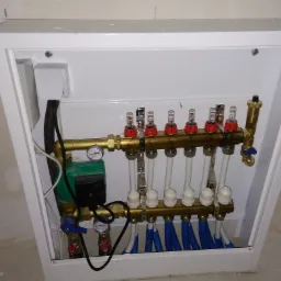 Rozdzielacz ogrzewania podłogowego w białej szafce, widoczne rury doprowadzające i odprowadzające wodę oraz pompa obiegowa.