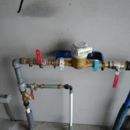 Zamontowany wodomierz z zaworami odcinającymi i niebieską rurą doprowadzającą wodę, widok na szarą ścianę i podłogę w tle.